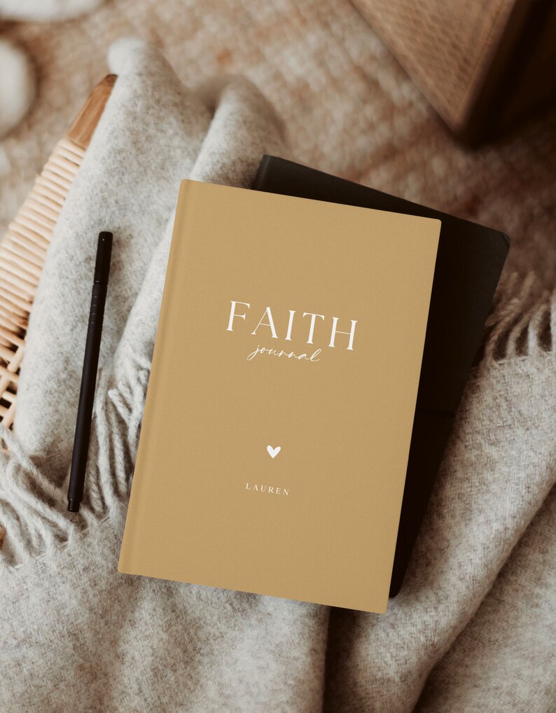 May include: A mustard-colored journal with the words "FAITH journal" in white script. A small white heart and the name "LAUREN" are below the text. A black pen rests on a gray blanket next to the journal. A second black journal is partially visible.