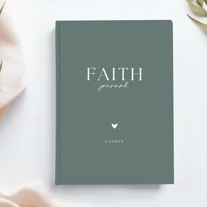 May include: A sage green journal with the words "FAITH journal" in white script. A small white heart and the name "LAUREN" are below the text. The journal is on a white surface with greenery and a pink cloth.