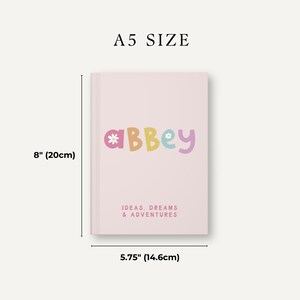 Może przedstawiać: R&oacute;żowy notes w formacie A5 z napisem "*aBbey*" w kolorowych literach z kwiatami na okładce. Notes zawiera napis "IDEAS, DREAMS & ADVENTURES" na dole. Notes ma wymiary 20 cm x 14,6 cm.