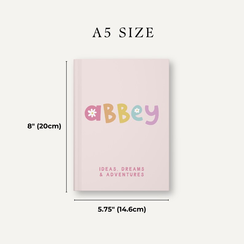 Może przedstawiać: R&oacute;żowy notes w formacie A5 z napisem "*aBbey*" w kolorowych literach z kwiatami na okładce. Notes zawiera napis "IDEAS, DREAMS & ADVENTURES" na dole. Notes ma wymiary 20 cm x 14,6 cm.