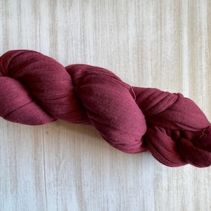 May include: A burgundy-colored scarf, knotted and twisted, with a fringed end. The scarf is made of a soft, woven material. The scarf is displayed on a light-colored wooden surface.