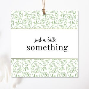 May include: Square white gift tag with a green paisley pattern border and the words "just a little something" in black script. The tag is suspended by a natural twine string.