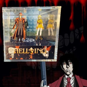 May include: A clear acrylic display box containing two anime character figures, one in a red coat and hat and one in a yellow uniform. The base features the Hellsing logo in gold text with a circular occult symbol. A character illustration is visible below.