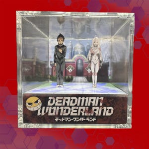 Peut inclure: Un présentoir en acrylique transparent avec deux personnages d'anime devant un arrière-plan détaillé. La base du présentoir affiche le texte "DEADMAN WONDERLAND" avec des caractères japonais. Le présentoir est posé sur un fond rouge.