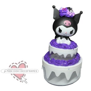 Puede incluir: Un pastel de dos niveles con un personaje negro con una calavera en la frente. El pastel tiene glaseado blanco con acentos de glaseado morado y chispas. El personaje tiene orejas negras y una cara blanca. Es un artículo decorativo.