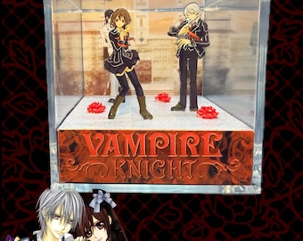 May include: A clear acrylic display case containing two anime-style character figurines standing on a white base with red rose accents. The front of the base features the text Vampire Knight in a stylized red font. A background image shows a building.