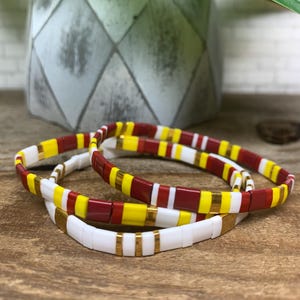 May include: Three beaded bracelets with a mix of colors. One bracelet is white with gold accents, another has red, yellow, and gold beads, and the third has red, white, and yellow beads. The bracelets are on a wooden surface.