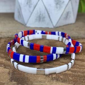May include: Three beaded bracelets in red, white, and blue. The bracelets are made of small, rectangular beads strung together with silver-colored metal accents. The bracelets are stacked on a wooden surface.