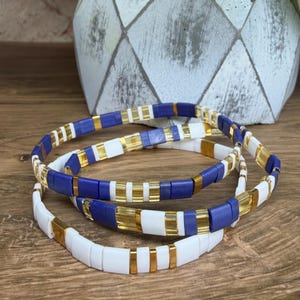 May include: Three beaded bracelets. Each bracelet features a repeating pattern of white, blue, and gold-colored rectangular beads. The bracelets are stacked on a wooden surface, with a decorative white and gray geometric planter in the background.