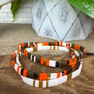 May include: Three beaded bracelets with a repeating pattern of white, orange, gold, and brown rectangular beads. The bracelets are stacked on a wooden surface. The bracelets are a stylish accessory.