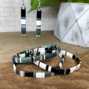 May include: A set of handmade jewelry featuring earrings and bracelets. The earrings are rectangular, with black, white, and teal segments. The bracelets have a similar design, with black, white, and teal rectangular beads. The jewelry is displayed on a wooden surface.