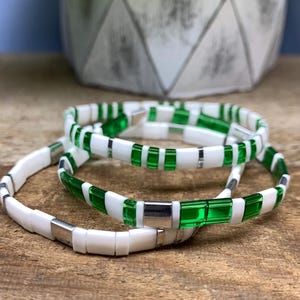 May include: Three beaded bracelets. The bracelets feature white and green rectangular beads with silver-colored metal clasps. The bracelets are stacked on a wooden surface, with a decorative vase in the background.