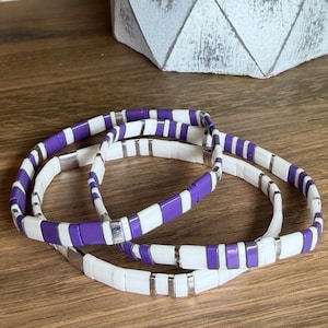 May include: Three stacked bracelets featuring a repeating pattern of rectangular purple and white beads separated by small, polished silver-toned metal spacers. The beads are arranged in a consistent sequence, creating a smooth, uniform appearance across each circular band.