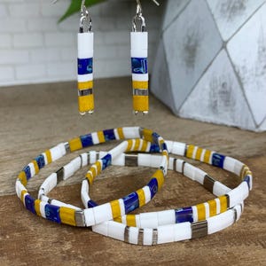 May include: A set of three bracelets and a pair of earrings made with white, blue, and yellow tiles. The bracelets are stacked on top of each other and the earrings are hanging from a silver stand.