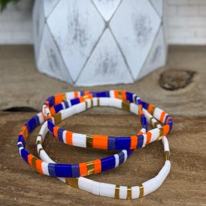 May include: Three beaded bracelets with a mix of white, orange, blue, and gold-colored accents. The bracelets are stacked on a wooden surface, with a white geometric planter in the background.