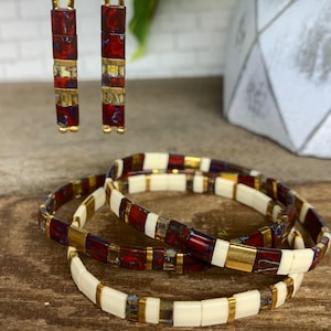 May include: Gold-toned earrings and bracelets. The earrings are rectangular with red, white, and gold accents. The bracelets have a similar design, featuring red, white, and gold rectangular beads. The jewelry is displayed on a wooden surface.