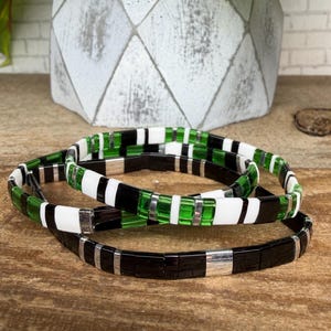 May include: Three beaded bracelets with a mix of black, white, and green rectangular beads. The bracelets have silver-toned metal accents and are displayed on a wooden surface. The bracelets are stacked on top of each other.
