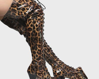 Op de afbeelding: Over-de-knie laarzen met een luipaardprint. De laarzen hebben een plateauzool en een stilettohak. Ze hebben een vetersluiting en een zijrits. De laarzen zijn gemaakt van een glanzend, lakachtig materiaal.