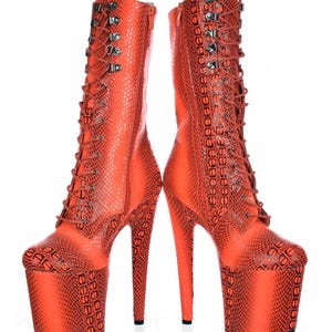 May include: A pair of bright orange platform boots with a snakeskin pattern. The boots have a lace-up design, a zipper on the side, and a stiletto heel. The boots are standing upright against a white background.