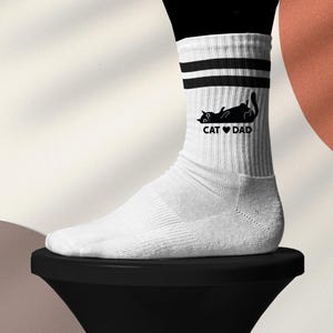 May include: White athletic socks with black stripes and a black cat graphic. The cat is lying down with the text "CAT ❤️ DAD" printed below. The socks are worn on a foot.