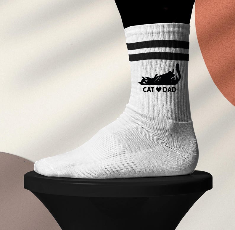 May include: White athletic socks with black stripes and a black cat graphic. The cat is lying down with the text "CAT ❤️ DAD" printed below. The socks are worn on a foot.