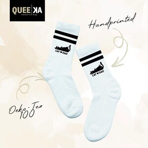 May include: A pair of white socks with black stripes at the top. Each sock features a black graphic of a cat and the words "CAT DAD". The socks are handprinted.