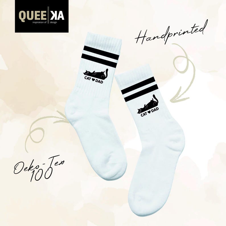 May include: A pair of white socks with black stripes at the top. Each sock features a black graphic of a cat and the words "CAT DAD". The socks are handprinted.