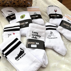 May include: A collection of white socks with various designs and text. Some socks feature black stripes, while others display graphics of an avocado, a wine glass, a cat, and the words "Lazy Sunday Club." Each pair is wrapped with a black band.