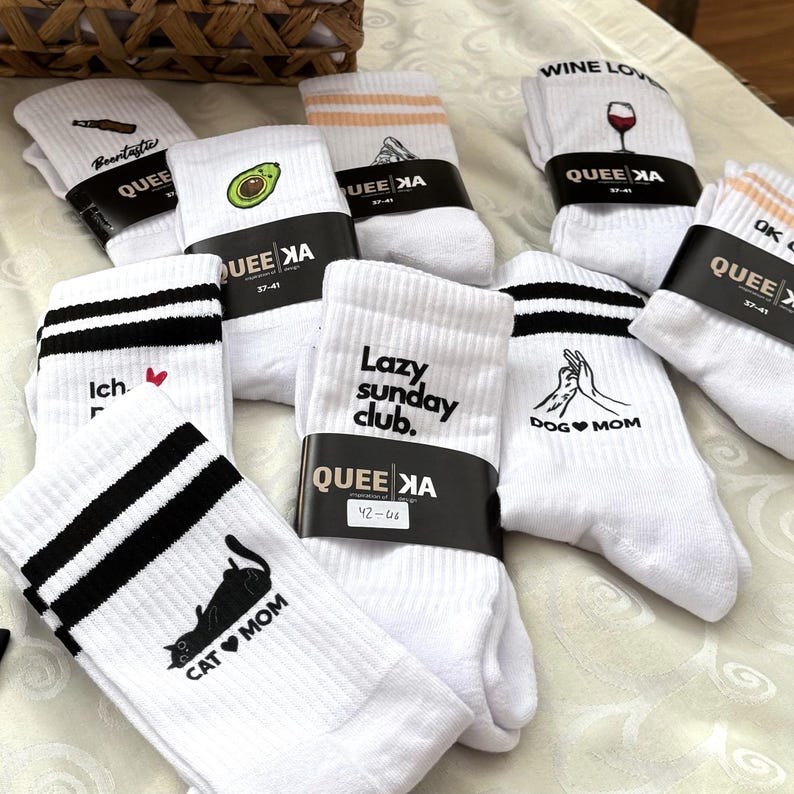 May include: A collection of white socks with various designs and text. Some socks feature black stripes, while others display graphics of an avocado, a wine glass, a cat, and the words "Lazy Sunday Club." Each pair is wrapped with a black band.