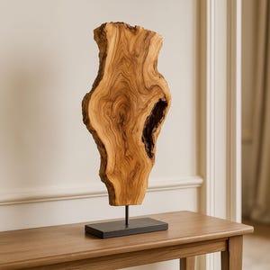 May include: A decorative wooden sculpture with a natural, organic shape. The light brown wood grain is visible throughout the piece, which is mounted on a dark rectangular base with a thin metal rod. The sculpture is displayed on a wooden table.