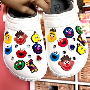 May include: White clogs decorated with colorful Sesame Street character charms. The charms include Elmo, Cookie Monster, Big Bird, and others. The clogs have a gray, fuzzy lining.