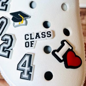 May include: White shoe with various decorative charms. Charms include the numbers "2024", a graduation cap, the words "CLASS OF", and an "I heart" charm. The charms are black, white, gray, and red.
