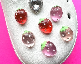 May include: White shoe with a rhinestone heart and multiple strawberry charms. The charms are in pink, red, and clear colors.