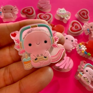 May include: A pink axolotl charm featuring a smiling face, wearing teal headphones, and holding a small game controller and a boba tea drink. The charm has a translucent border and a small red heart accent on the side, showcasing a detailed design.