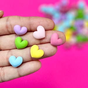May include: Six small heart-shaped decorations in various pastel colors, including pink, light blue, yellow, green, and lavender. The hearts are arranged on a hand.