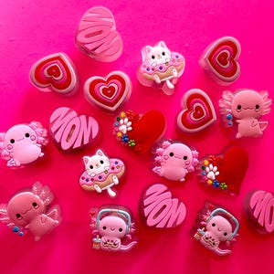 May include: A collection of small shoe charms featuring various designs including pink hearts with the word MOM, red hearts with colorful paw prints, white cats on donuts, and pink axolotls, some wearing headphones or holding drinks, scattered on a surface.