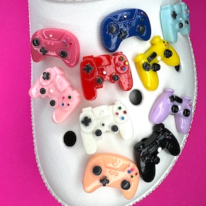 May include: A set of ten colorful plastic video game controller charms. The charms are in various colors, including pink, red, yellow, blue, white, black, and purple. The charms are all in the shape of a video game controller.