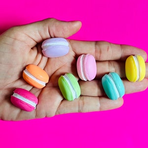 May include: A hand holding eight colorful macarons. The macarons are in shades of pink, orange, yellow, green, and purple.