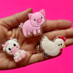 May include: Three farm animal charms: a pink pig, a white and pink cow, and a white chicken with a red comb and yellow beak. The charms are on a bright pink background.