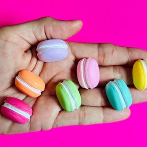 May include: A hand holding seven colorful macarons. The macarons are in shades of pink, purple, yellow, green, and orange.