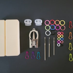 May include: A set of sewing tools and accessories in a white case. The set includes a pair of small scissors, a needle threader, needles, and colorful plastic rings.