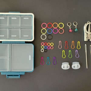 May include: A blue and white plastic storage box with multiple compartments, filled with colorful stitch markers, rings, and needles. The box also includes a pair of small scissors, two needle threaders, and a small bell charm.