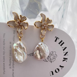 May include: A pair of gold earrings with white flower charms and iridescent pearl drops. The earrings are displayed on a card with the text "THANK YOU".