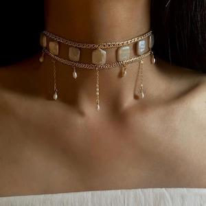 May include: A gold-toned choker necklace featuring two delicate chain layers connected by five rectangular, iridescent white flat beads. Several small, teardrop-shaped white pearls hang from the lower chain on thin gold-toned metal links.