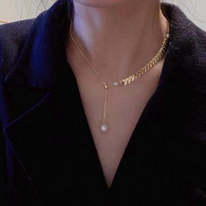 May include: A gold-toned necklace featuring a delicate chain with a series of small, leaf-shaped metal links on one side. The necklace includes a single white pearl accent at the junction and a longer chain segment ending in a dangling white pearl.