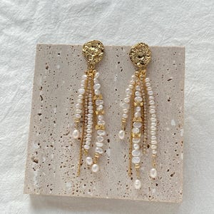 Peut inclure: Boucles d'oreilles dorées avec un dessus circulaire texturé. Les boucles d'oreilles présentent plusieurs brins de petites perles blanches et des accents dorés, créant un effet pendant. Les boucles d'oreilles sont présentées sur une surface beige clair texturée.