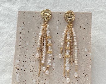 May include: A pair of gold-tone drop earrings featuring textured circular studs. Each earring has multiple dangling strands composed of small white freshwater pearls, thin gold-tone chains, and delicate gold-tone bow accents.