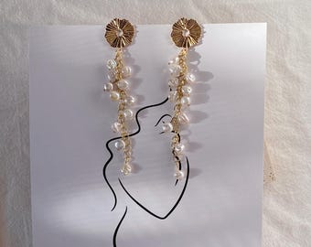 May include: Gold-toned dangle earrings featuring a floral stud design and cascading white pearls. The earrings hang from a gold-colored post and are displayed on a white background with a minimalist line drawing.