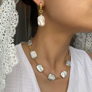 May include: A gold-toned hoop earring and a necklace with large, irregular-shaped white pearls. The necklace is strung on a gold-colored chain. The jewelry is worn with a white top.