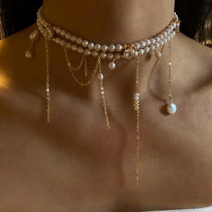 May include: A double-strand pearl choker necklace featuring gold-tone chains, dangling pearl accents, and a central clear round gemstone. The design includes multiple delicate gold chains hanging from the pearl strands at varying lengths.